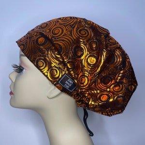 May include: A black scrub hat with a gold and black swirl pattern. The hat has a black drawstring and a small black label that says "ANIA MADE THIS".