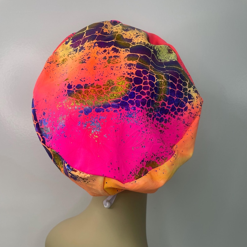 May include: A pink satin bonnet with a colorful abstract print featuring a snakeskin pattern. The bonnet has a tie string.