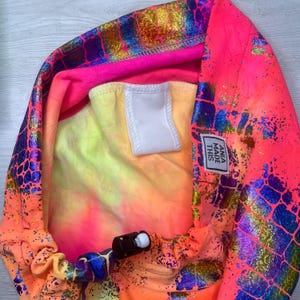 May include: A pink, yellow, and blue tie-dye fabric with a white label that says "ANIA MADE THIS". The fabric has a holographic, iridescent, and scaly pattern.
