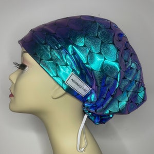May include: A turquoise and purple scrub hat with a mermaid scale pattern and a white drawstring. The hat has a label that says "Aniamadethis".