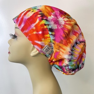 May include: A colorful tie-dye scrub cap in shades of orange, pink, yellow, and blue. The cap features a tag that reads "ANIA MADE THIS". The cap is designed to be worn on the head.