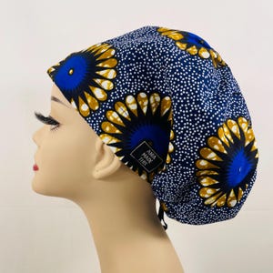 May include: A blue and gold patterned scrub cap with white polka dots. The cap features a floral design and a small tag that reads "ANIA MADE THIS". The cap is worn by a mannequin.