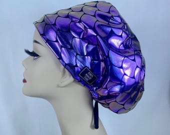 May include: A metallic purple scrub cap featuring a repeating black scale pattern. The fabric has a shiny, iridescent finish. A small black rectangular tag on the side displays the white text ANIA MADE THIS. The cap includes a black tie closure.