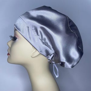 May include: A silver satin bonnet with a drawstring closure. The bonnet is designed to protect hair and maintain hairstyles. The fabric has a subtle sheen, and the drawstring is white. The bonnet is shown on a mannequin head.