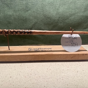 May include: A handcrafted wooden wand made of red oak, 11.5 inches long, displayed on a wooden stand. The wand has intricate carved details and is accompanied by a tag. The stand has the text "GordBeams Shop" on it.