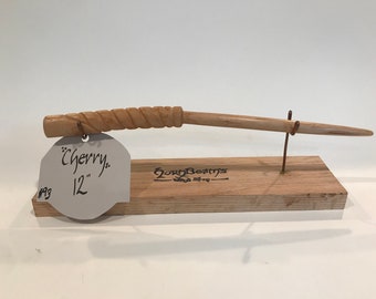 Op de afbeelding: Een 30,5 cm lange houten toverstaf gemaakt van kersenhout met een gedraaid ontwerp. De toverstaf wordt getoond op een houten standaard met een metalen draadhouder. Het label op de standaard luidt "Cherry" en "12" en "GoryBeams Wood Shop".