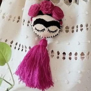 May include: A handmade Frida Kahlo-inspired pendant. The pendant features a white face with black eyebrows and eyelashes, adorned with pink felt roses and a matching tassel. The background is a white, textured fabric.