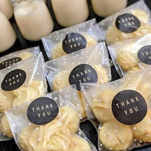 May include: An overhead shot shows small glass jars filled with a creamy liquid, alongside clear cellophane bags of cookies. Each bag is sealed with a black circular sticker that reads "THANK YOU" in white lettering. The cookies are a light yellow color.