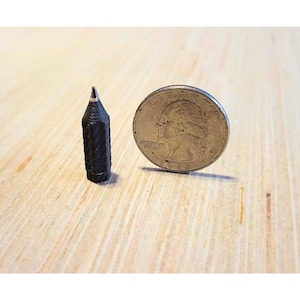 May include: A miniature black pencil next to a quarter coin. The pencil is small, with a hexagonal body and a pointed tip. The quarter coin shows a profile of George Washington.