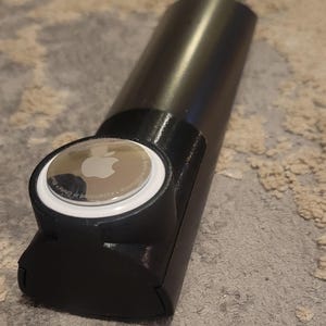 May include: Black cylindrical object with a rounded end, holding a silver Apple AirTag. The AirTag is white with a silver Apple logo. The object appears to be a holder or case for the AirTag.