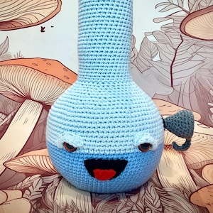 May include: A light blue crochet bong with a smiling face, red mouth, and brown eyes. The bong has a rounded base and a long, cylindrical neck. A small, gray crochet piece is attached to the side. The background features a mushroom-themed design.
