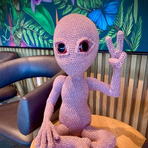 Puede incluir: Una figura extraterrestre de crochet con grandes ojos rojos, sentada con las piernas cruzadas y haciendo un signo de paz con una mano. La figura es de color rosa claro y está sentada en una mesa de madera.