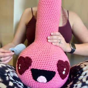 Könnte beinhalten: Ein rosa gehäkeltes Plüschtier in Form einer Comicfigur mit einem lächelnden Gesicht, schwarzen Augen und zwei roten Herzen als Pupillen. Das Spielzeug hat einen rosa Mund und eine schwarze Nase.