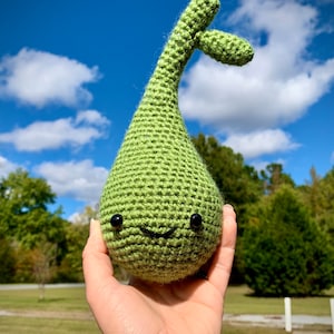 May include: A green crocheted plush toy shaped like a teardrop with a small stem and a smiling face with black eyes. The toy is being held in a person's hand against a blue sky background.