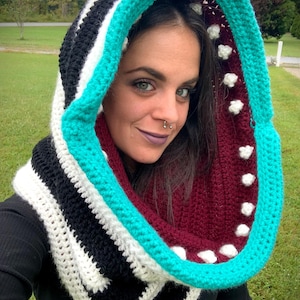 May include: A crocheted cowl with a large hood. The cowl is made with black, white, and turquoise yarn. The hood has a burgundy lining with white polka dots.