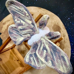 May include: A crocheted moth with white, gray, and purple wings. The moth is sitting on a wooden stool in front of a backdrop of a moon and stars.