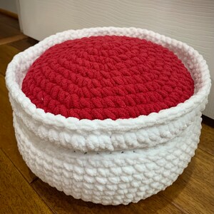 May include: A white crocheted basket with a red crocheted cushion inside. The basket is round and has a soft, fluffy texture.