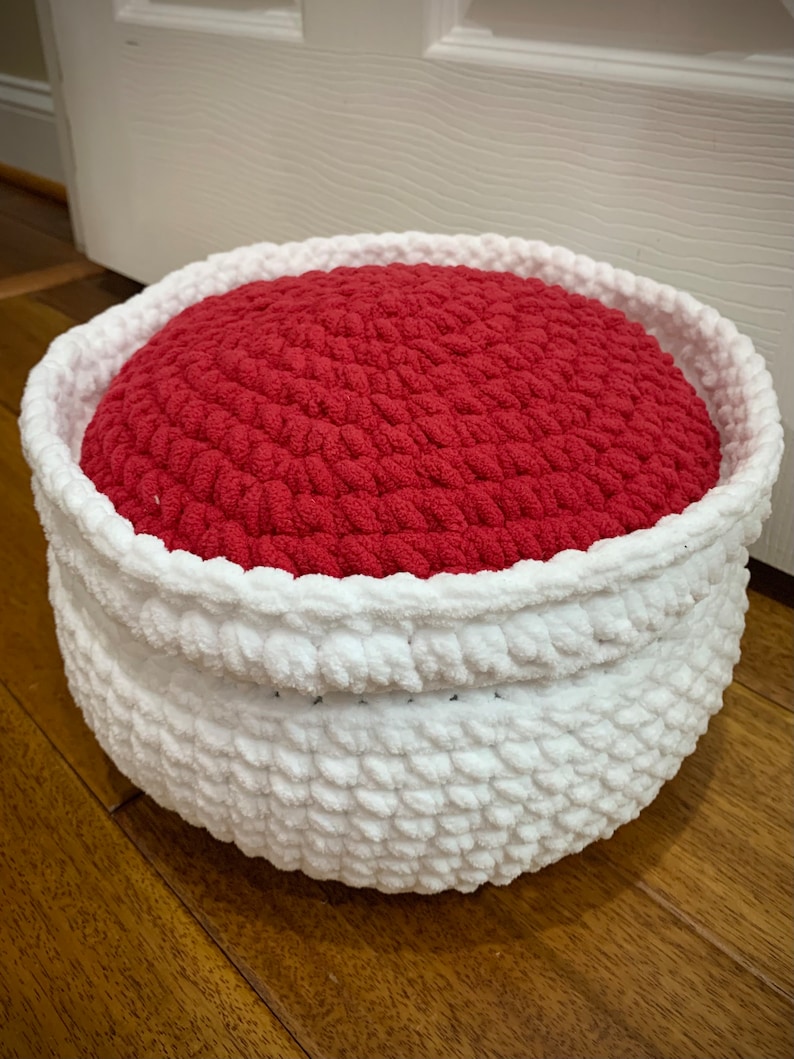 May include: A white crocheted basket with a red crocheted cushion inside. The basket is round and has a soft, fluffy texture.