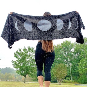 May include: A black knit blanket with a white moon phase design. The blanket is being held up by a person in a field.