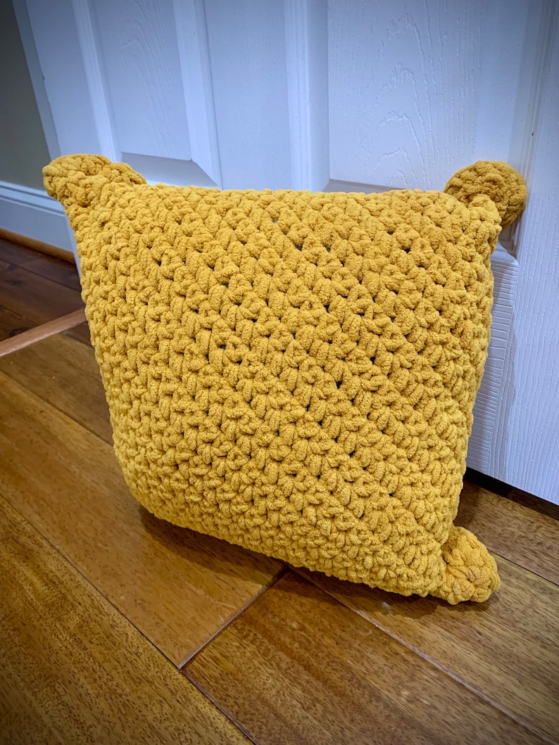 May include: A mustard yellow crocheted cushion with pom-poms on the corners.