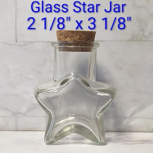 May include: Clear glass star-shaped jar with a cork stopper. The jar is 2 1/8" x 3 1/8" and has the words "Glass Star Jar" in blue text. The jar is empty and sits on a white surface.
