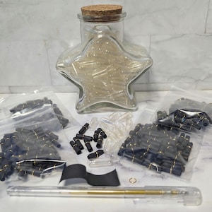 May include: A clear star-shaped glass jar filled with capsules, a clear pen, and several small bags containing black and gold components. A cork-topped jar is also visible. The items are arranged on a white surface.