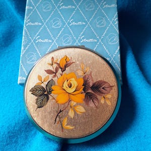 May include: A vintage Stratton compact mirror with a gold finish. The compact has a floral design with a yellow rose and brown leaves. The light blue box has the word "Stratton" printed on it.