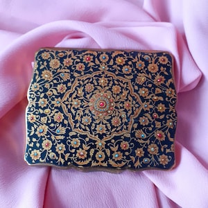 May include: A black and gold vintage compact with a floral design and colorful jewels. The compact has a detailed intricate pattern.