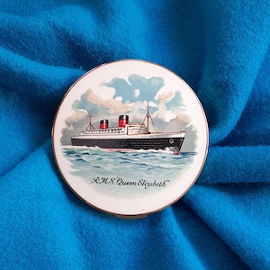 May include: A circular vintage compact featuring a detailed illustration of the RMS Queen Elizabeth ocean liner. The image shows the ship sailing on blue waves under a cloudy sky. The compact has a gold-coloured rim and the text "R.M.S. Queen Elizabeth".