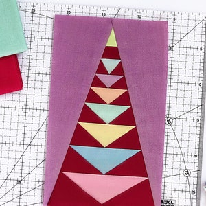 May include: A close-up of a quilt block with a purple background and a red, pink, and blue triangle pattern. The block is on a white cutting mat with a grid pattern. There are also spools of thread and a rotary cutter in the image.