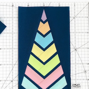 May include: A close-up of a quilt block featuring a colourful geometric design. The block is made up of seven triangles in a pyramid shape, with the bottom triangle being the largest and the top triangle being the smallest. The triangles are in shades of pink, blue, green, and yellow. The block is on a blue background and is surrounded by a white grid.