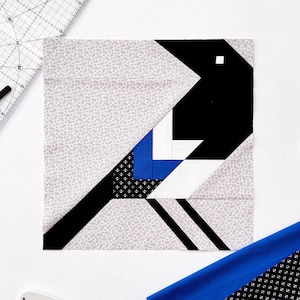 May include: A geometric fabric block featuring a black and white magpie design. The bird's body is black, with white and blue accents. The block is surrounded by quilting tools, including a ruler, scissors, and fabric.