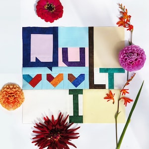 Op de afbeelding: Een kleurrijk quiltblok met het woord "QUILT" gespeld in verschillende gekleurde stoffen. Het blok is omringd door bloemen in tinten rood, oranje en roze.