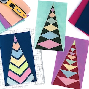May include: Three fabric Christmas tree quilt blocks in various colors, including pink, blue, green, yellow, and red. The blocks are on a white surface with a ruler, rotary cutter, and spools of thread.