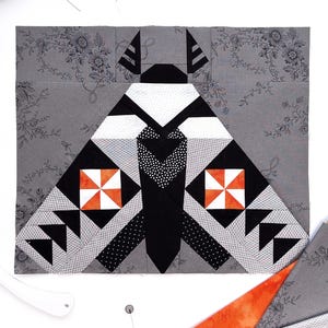 May include: A square quilt block featuring a moth design constructed from fabric pieces in shades of gray, black, white, and orange. The moth has patterned wings with geometric shapes, including orange and white pinwheel squares and black triangles.