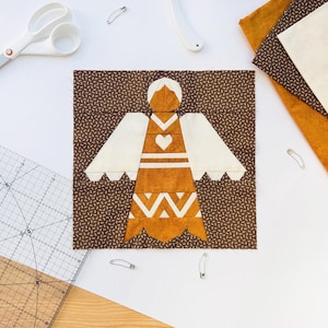 May include: A square quilt block featuring a central angel figure pieced from brown, mustard yellow, and cream fabric. The angel has a heart detail on the chest and geometric patterns on the skirt, set against a dark brown background with small white dashes.