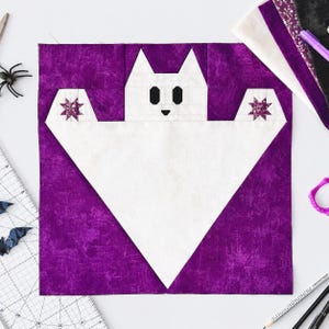 Pode incluir: Um bloco de colcha quadrado apresentando uma forma de fantasma branco centralizada em um fundo roxo texturizado. O fantasma tem dois olhos pretos, um pequeno nariz preto e dois pequenos detalhes em forma de estrela roxa nos braços estendidos.