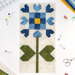 May include: A fabric flower quilt block with a blue and yellow center, surrounded by blue heart-shaped petals. The stem and leaves are green. The background is a light beige. Sewing notions, including pins and scissors, are scattered around the block.