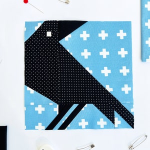 May include: A black crow fabric block with white polka dots on a blue background with white plus signs. The crow is made up of several fabric pieces.