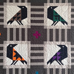 May include: A quilt with a black and white striped background and four black birds with colorful accents. The birds are arranged in a grid pattern with a purple flower in the center of the top row and a blue flower in the center of the bottom row.
