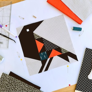 Op de afbeelding: Een vierkant quiltblok met een gestileerd vogelontwerp, samengesteld uit stofresten in effen zwart, feloranje en patronen in grijs, zwart en blauwgroen, omringd door naaibenodigdheden zoals een liniaal, spelden en garen.