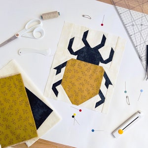 May include: A close-up of a quilt block featuring a black and white geometric design of a beetle with a yellow hexagon body. The block is surrounded by sewing tools, including scissors, thread, pins, and a ruler.