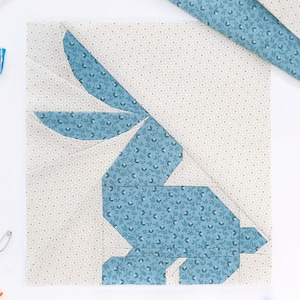 May include: A square fabric block featuring a blue and white bunny design. The bunny is constructed from blue floral patterned fabric, set against a cream background with a small floral pattern. The design is a patchwork or quilting project.