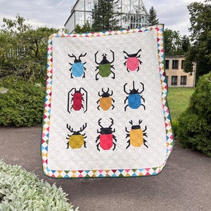 Pode incluir: Um edredom branco com um padrão repetido de besouros coloridos e estilizados. O edredom tem uma borda geométrica com uma variedade de cores. O fundo inclui uma estufa e vegetação exuberante.