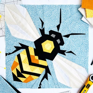 May include: A colorful, geometric bee quilt block. The bee features a black body with yellow and orange accents, set against a light blue floral background. White and patterned wings extend from the bee. Sewing tools and fabric scraps surround the block.