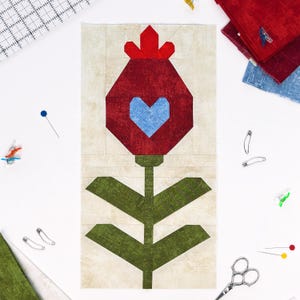 May include: A fabric quilt block featuring a stylized red flower with a blue heart center and green stem and leaves. The block is surrounded by sewing supplies, including pins, safety pins, and scissors, on a white surface.