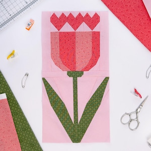 Pode incluir: Um bloco de tecido de tulipa em tons de rosa, vermelho e verde, com um fundo rosa claro. A tulipa tem um design detalhado, com um caule e folhas verdes. A imagem é um close-up, mostrando os detalhes do acolchoado.
