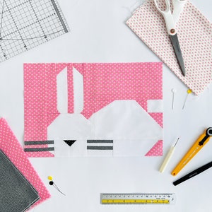 May include: A pink and white fabric bunny applique with black eyes and nose. The bunny is surrounded by sewing tools, including a ruler, scissors, pins, and a rotary cutter.