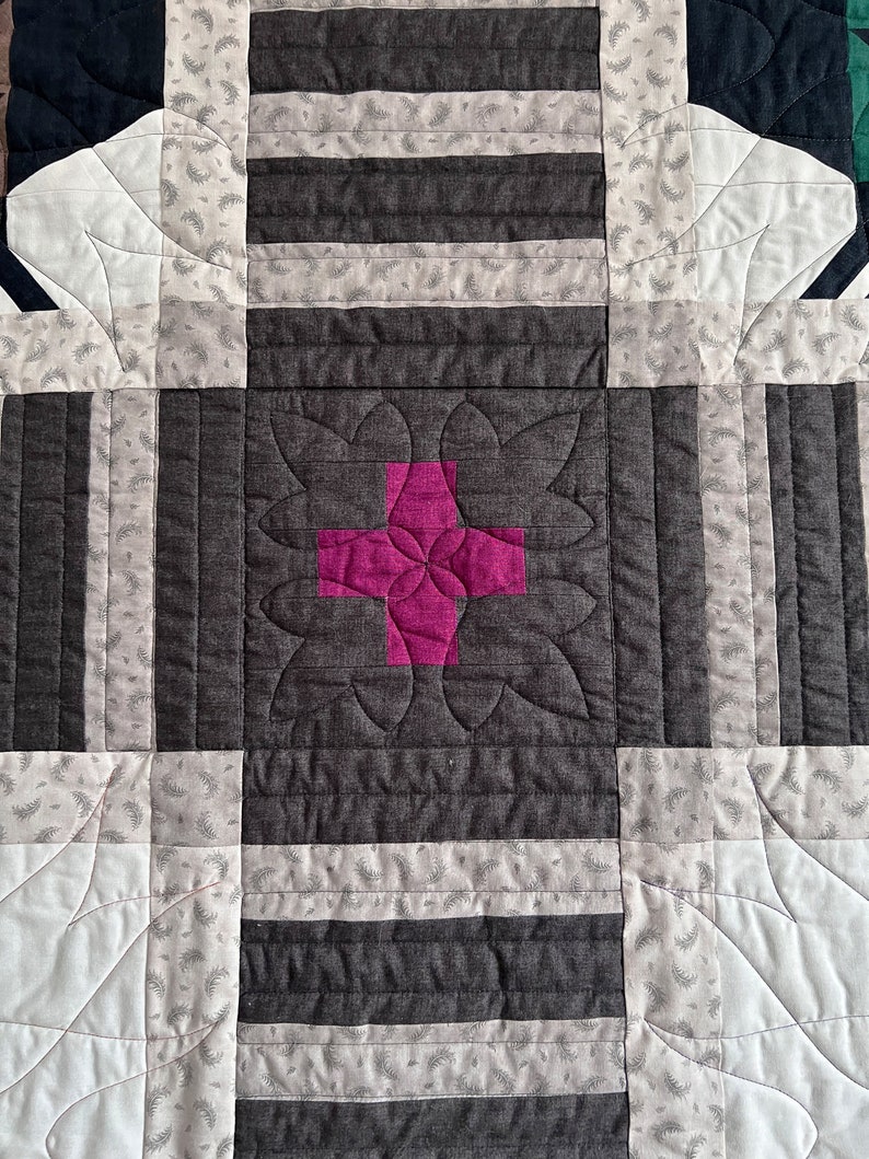 May include: A quilt block with a pink plus sign in the center, surrounded by a floral design. The block is made of various fabrics, including a striped fabric and a fabric with a small leaf pattern.