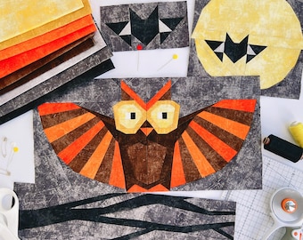 May include: A collection of fabric quilt blocks featuring an owl with orange and brown wings, a bat silhouette against a yellow moon, and a small bat block. Includes a stack of yellow, orange, and brown fabric squares, sewing pins, and a rotary cutter.